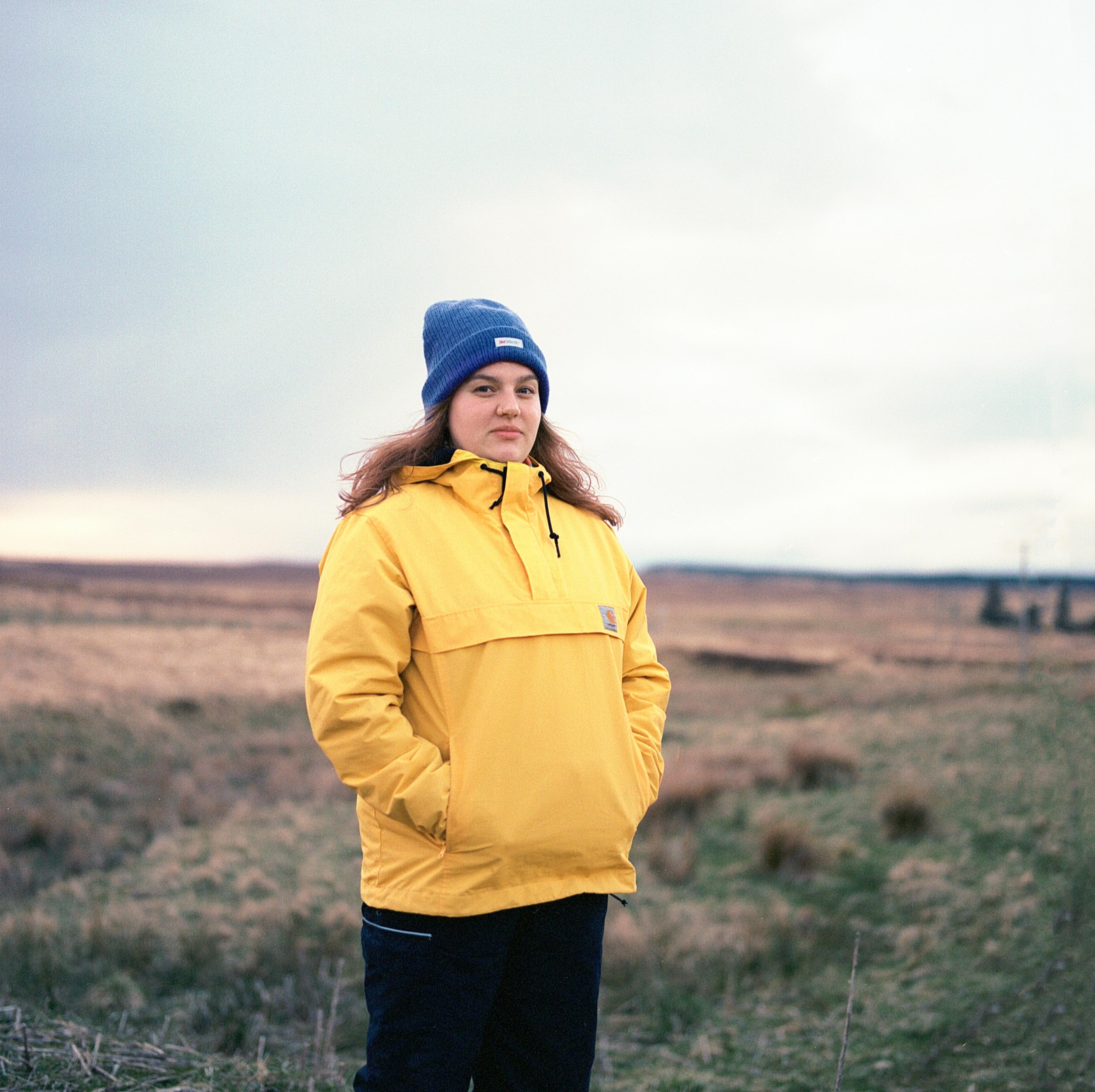 Molly standing in a wide open landscape, looking into the camera. She is wearing a bright yellow coat and purple woolly hat.