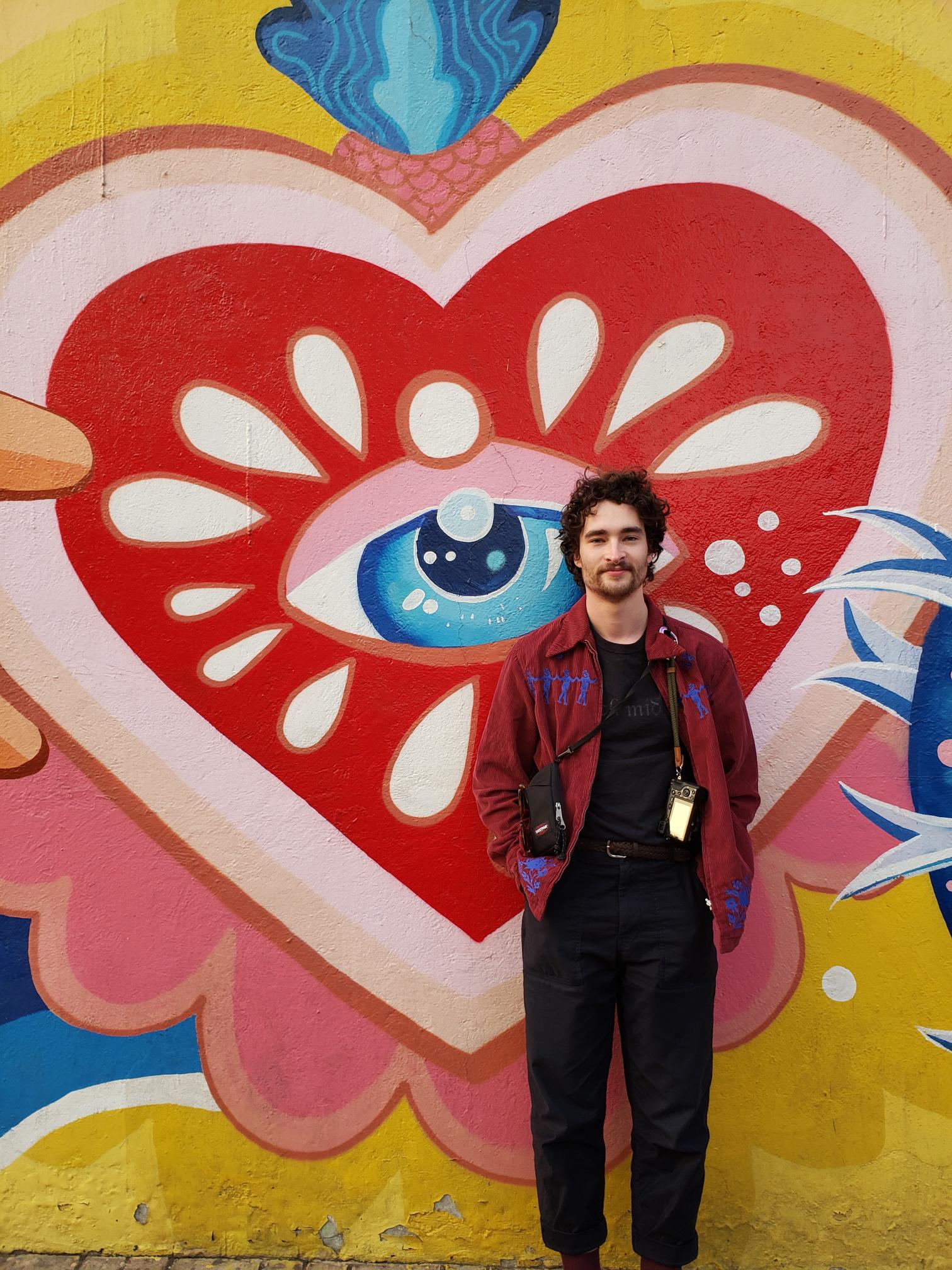 Sam stadning against a wall which has a brightly colored eye and heart painted on it. He is wearing a read shirt and has a camera over his shoulder.
