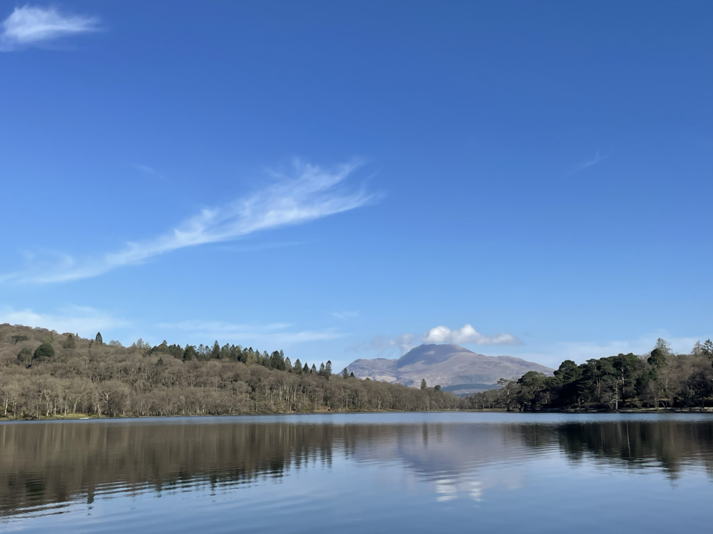 A view over Loch Lomond with hills and trees in the background. The waters is still and the sky is blue.